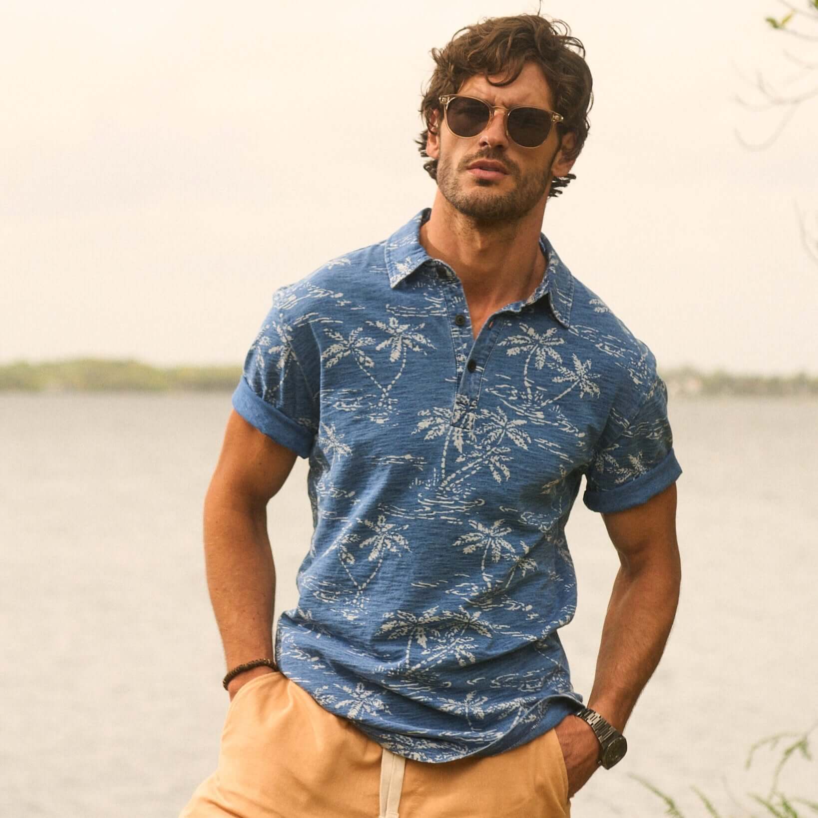 Man wearing a short sleeve dark indigo polo shirt with a tropical print, styled casually by the water.