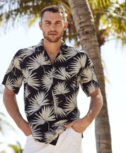 Short Sleeve Rayon Print Camp Collar In Sky Captain modeled by a man in a tropical setting with palm trees.