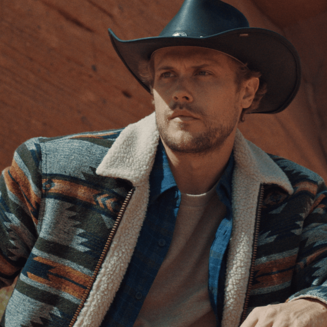 Man wearing a southwestern sherpa-lined jacket and cowboy hat, posing against a rustic background.