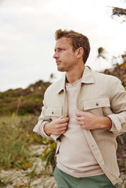 Man in a beige jacket and light sweater poses outdoors on the beach, with greenery and a cloudy sky in the background.