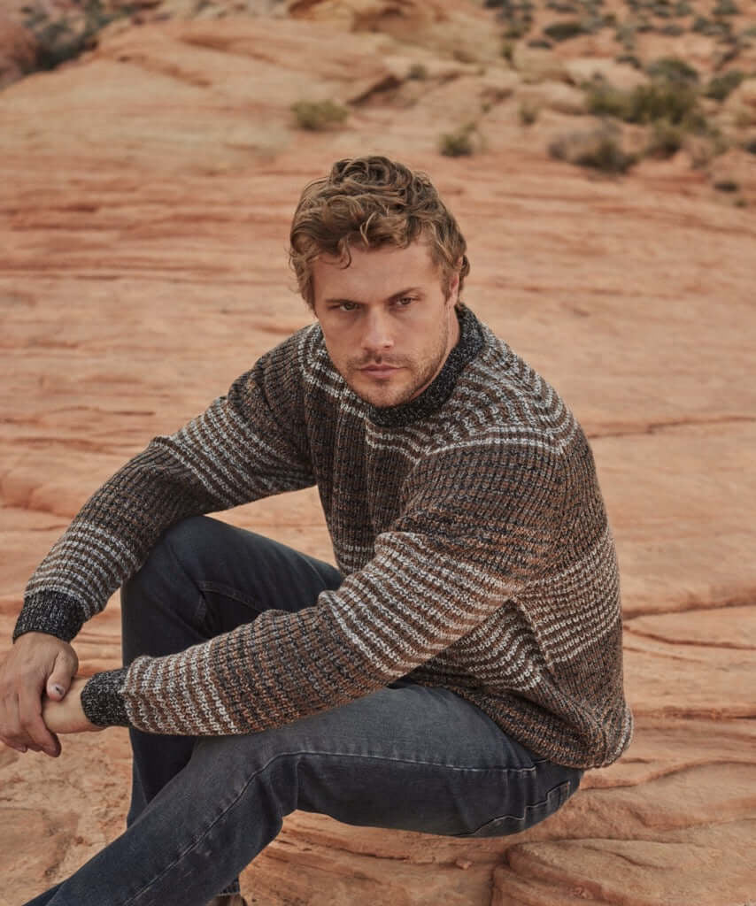 Model wearing a Tonal Stripe Crew in Natural Mix, featuring a 100% acrylic fabric, seated on rocky terrain.