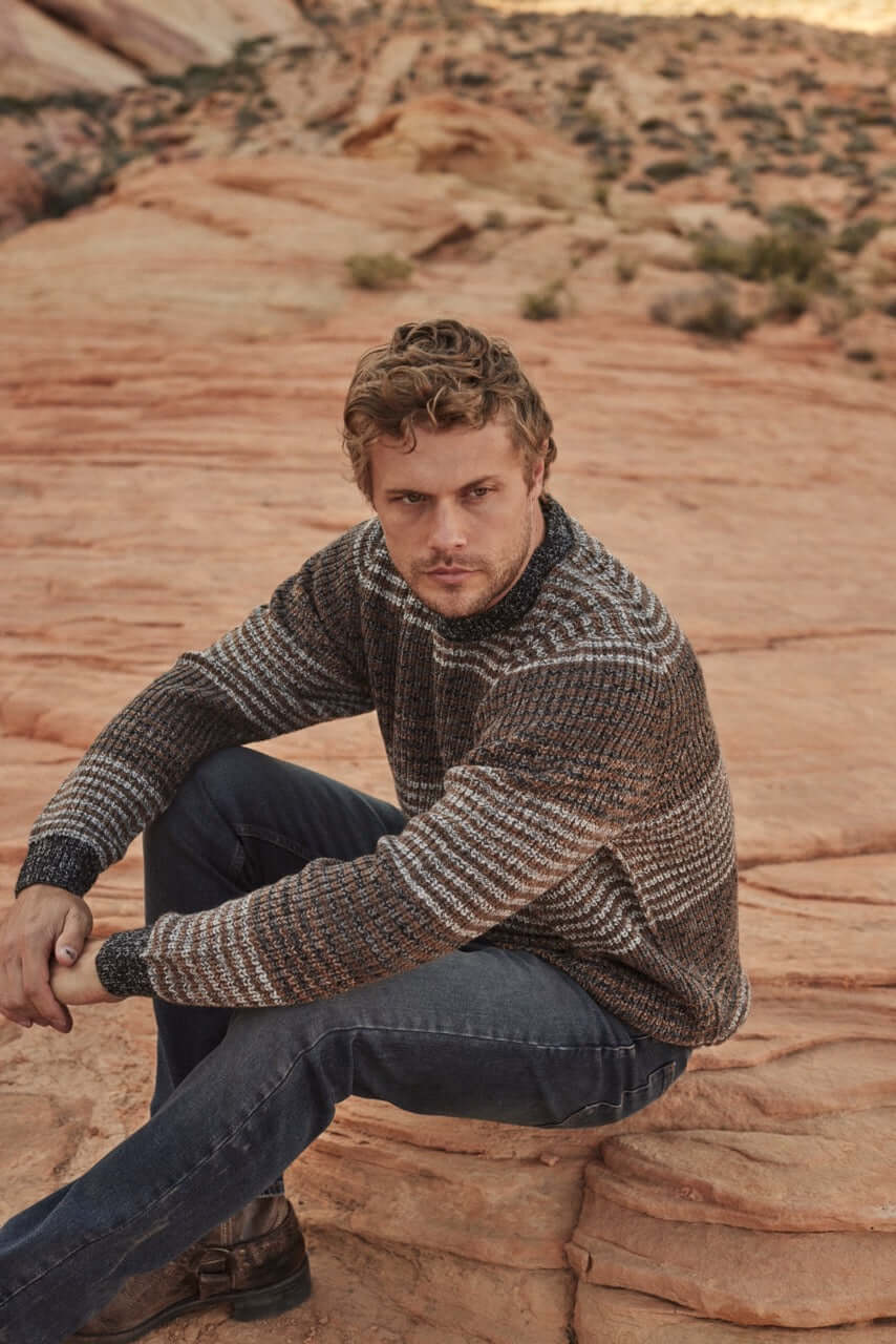 Model wearing a Tonal Stripe Crew in Natural Mix, featuring a 100% acrylic fabric, seated on rocky terrain.