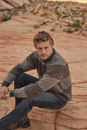 Model wearing a Tonal Stripe Crew in Natural Mix, featuring a 100% acrylic fabric, seated on rocky terrain.