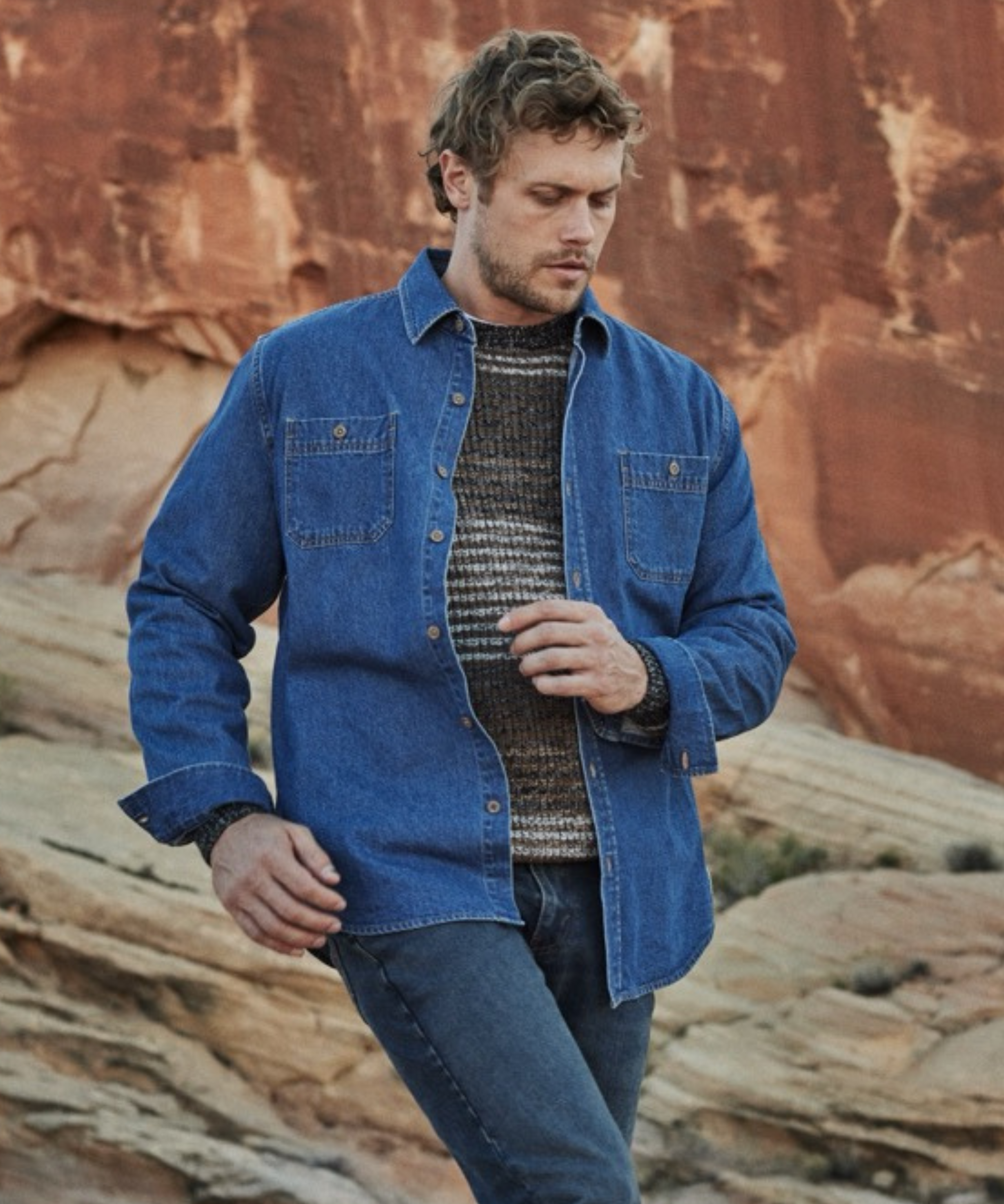 Man wearing unlined medium indigo denim overshirt over a knit sweater, set against a rugged natural landscape.