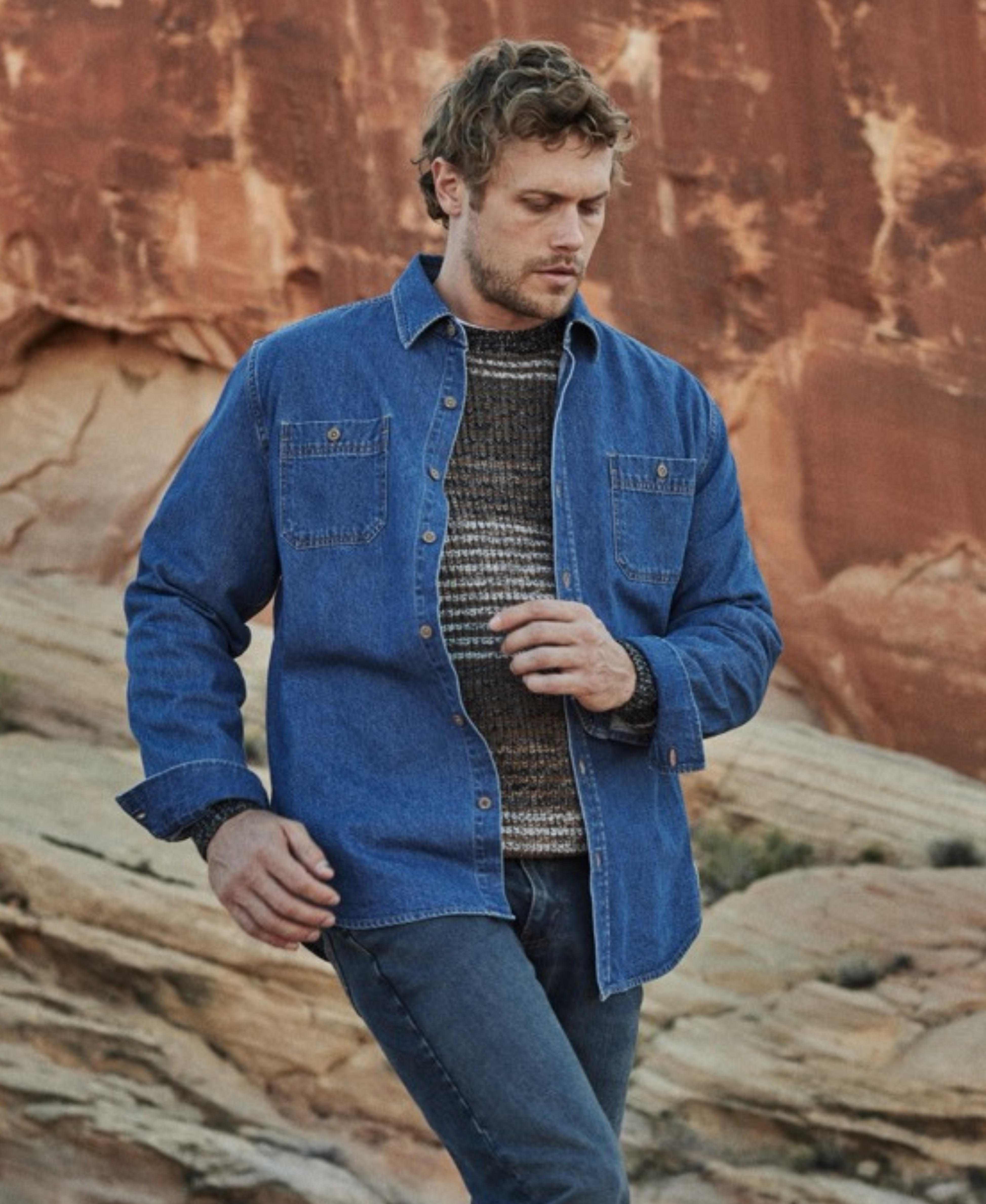 Man wearing unlined medium indigo denim overshirt over a knit sweater, set against a rugged natural landscape.