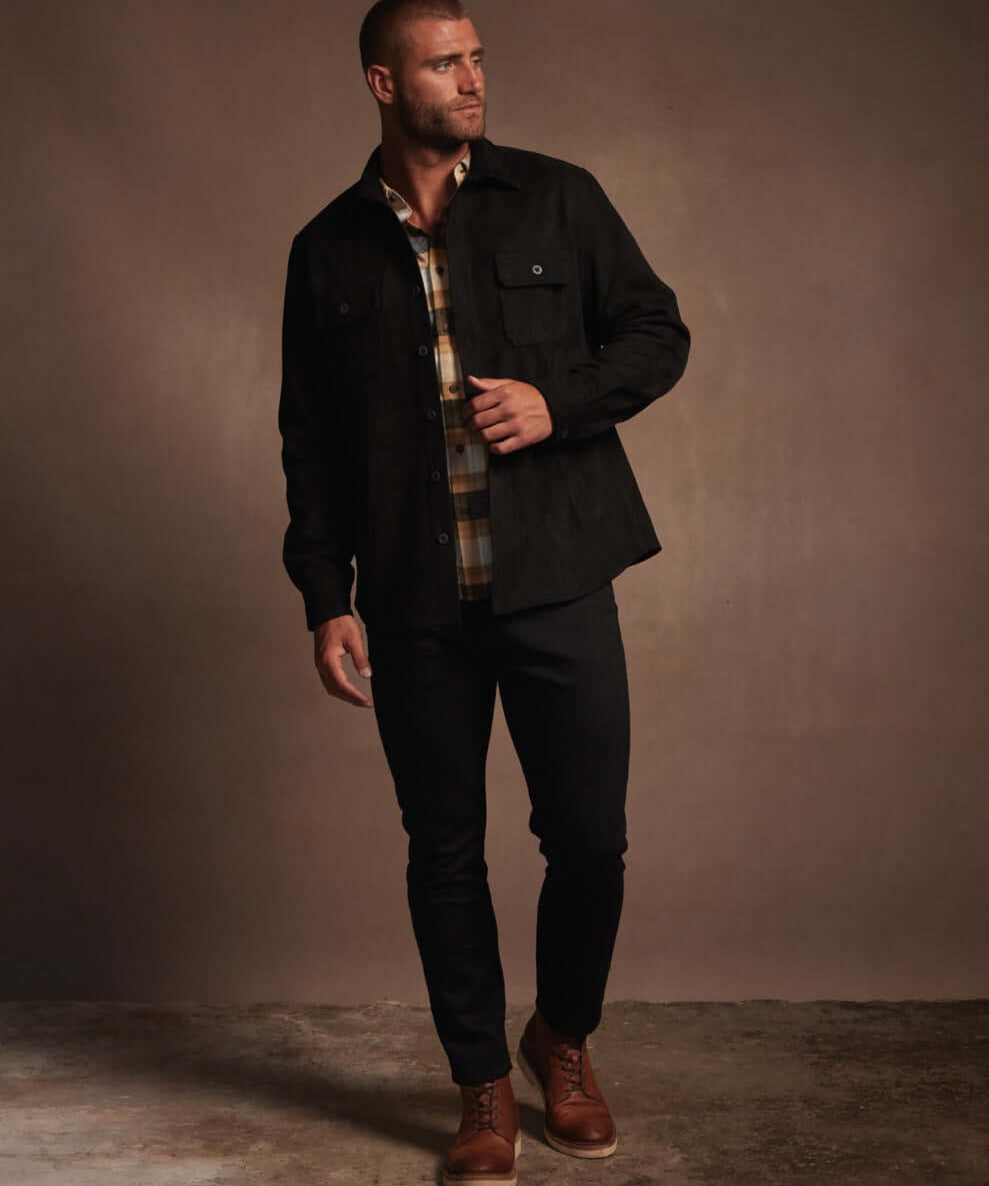Model wearing an unlined sueded shirt jacket in black, featuring side pockets, styled with plaid shirt and dark pants.