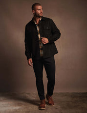Model wearing an unlined sueded shirt jacket in black, featuring side pockets, styled with plaid shirt and dark pants.