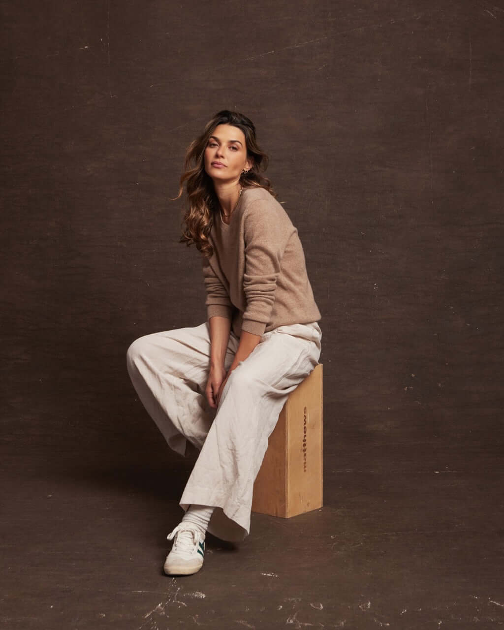 Woman wearing a taupe heather cashmere sweater paired with cream wide-leg pants, seated on a wooden block.