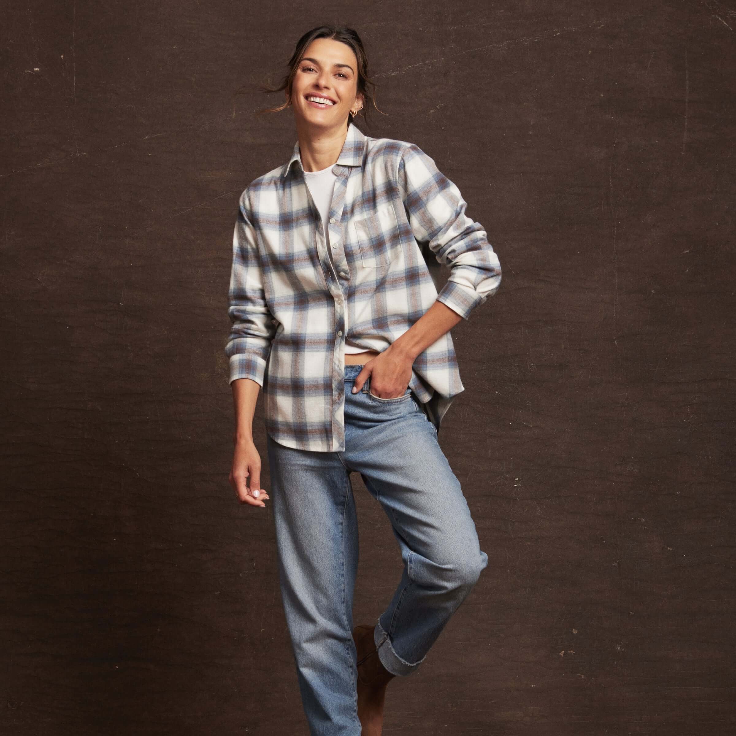 Smiling woman wearing a blue checkered women's stretch flannel shirt with rolled sleeves and relaxed jeans.
