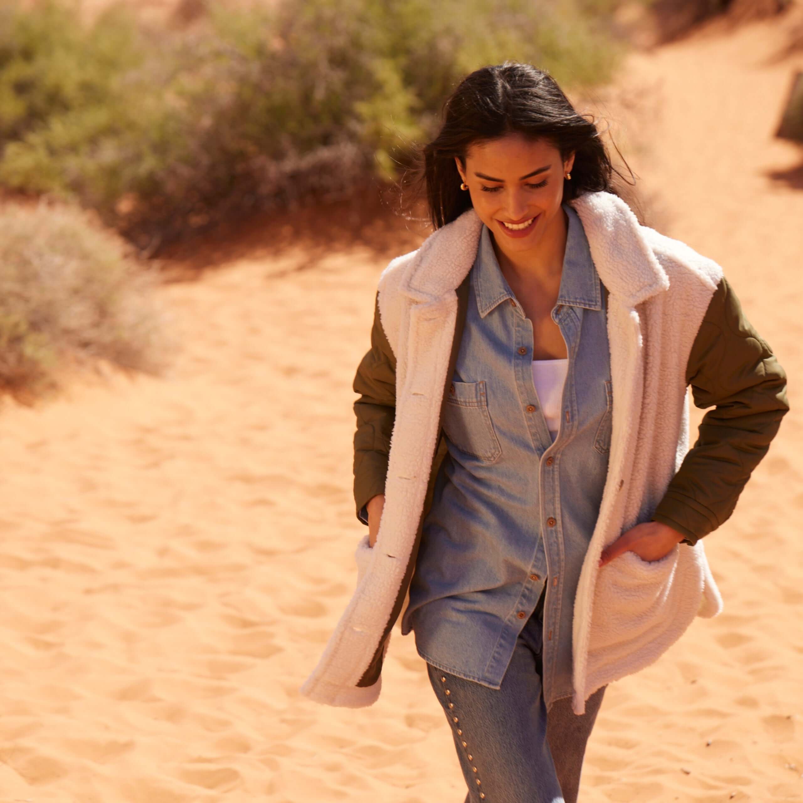 Smiling woman in a cozy women's quilted mixed media jacket, walking on sandy terrain, showcasing casual outdoor style.